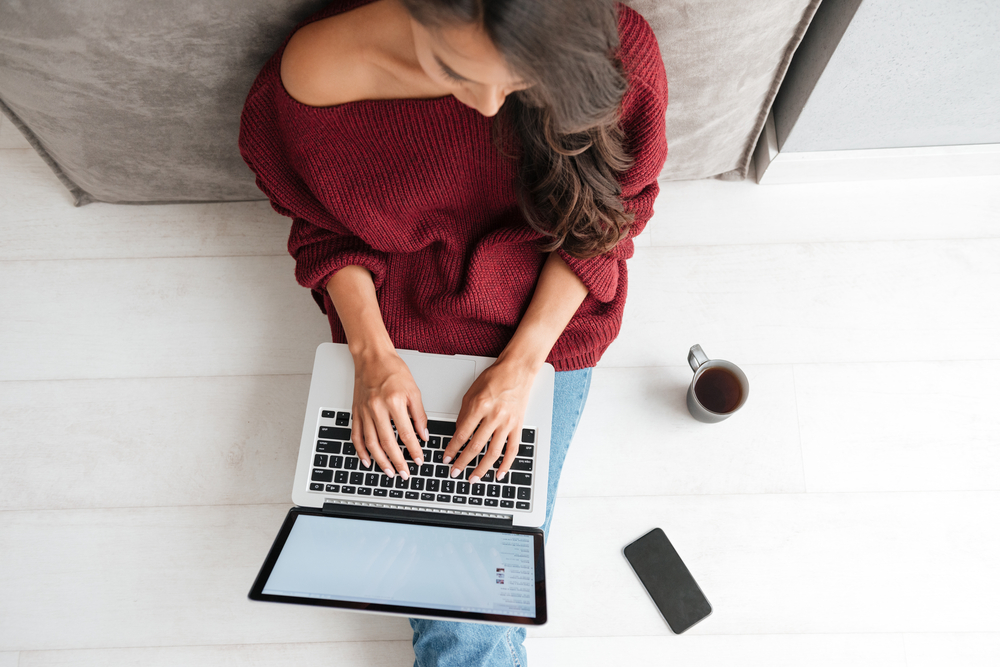A-professional-woman-with-brown-hair-and-a-maroon-sweater-works-on-her-laptop-computer-while-sitting-on-a-white-floor,-with-a-cup-of-coffee-next-to-her