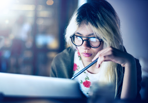 A-blonde-woman-wearing-glasses-studies-at-her-laptop-while-holding-a-pen-in-her-hand.
